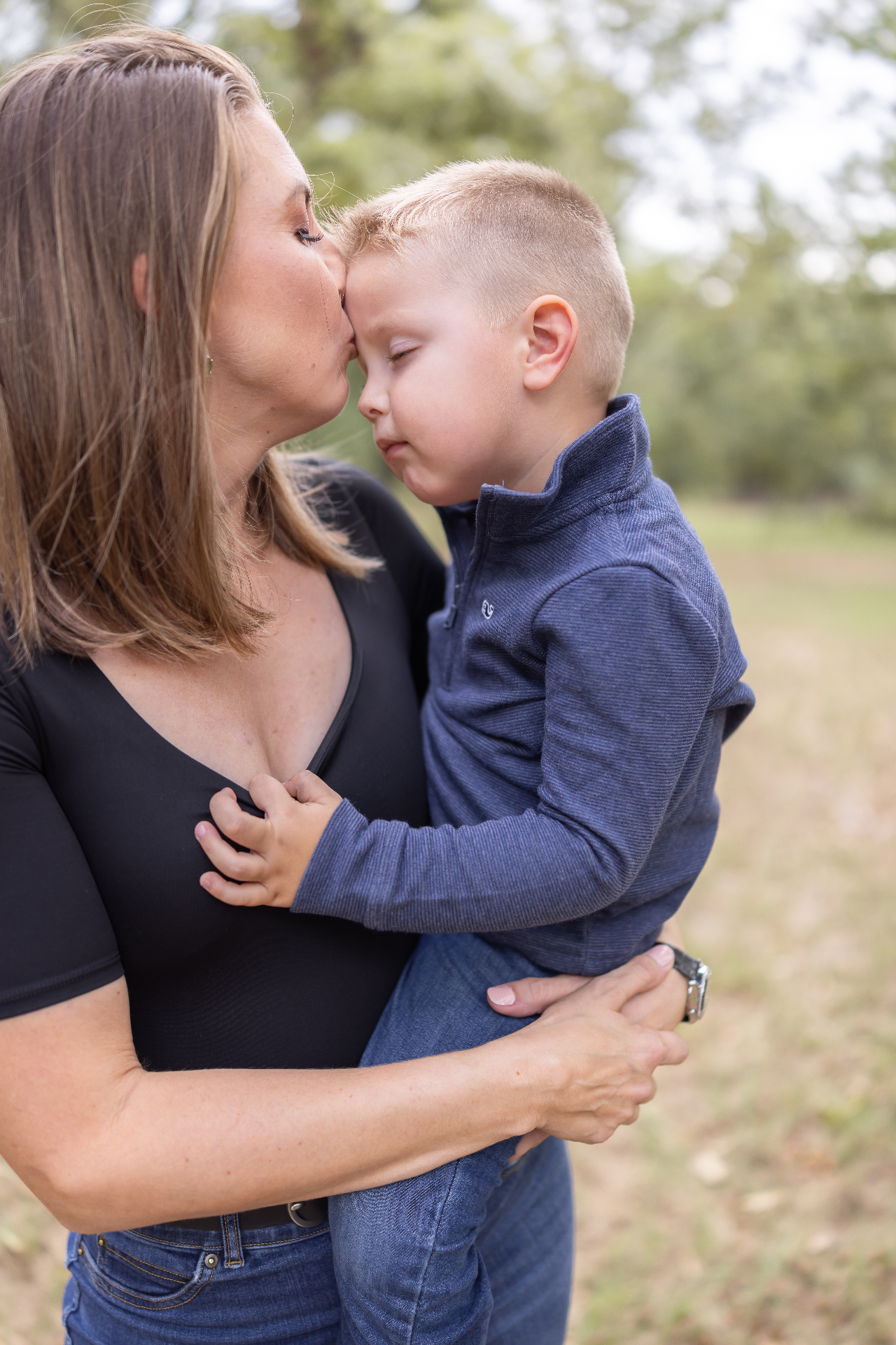 A mother tenderly kisses her young son's forehead outdoors — a quiet moment of calm and connection
